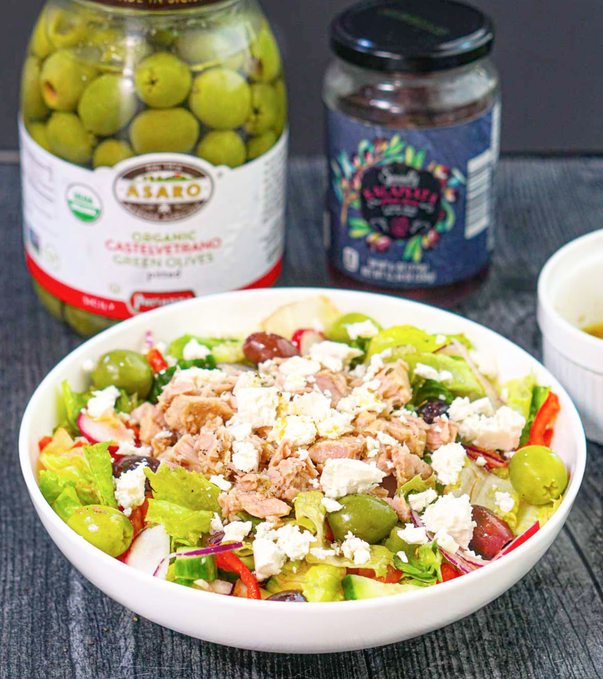 White bowl with the Greek flavored salad and jars of different olives in the background.