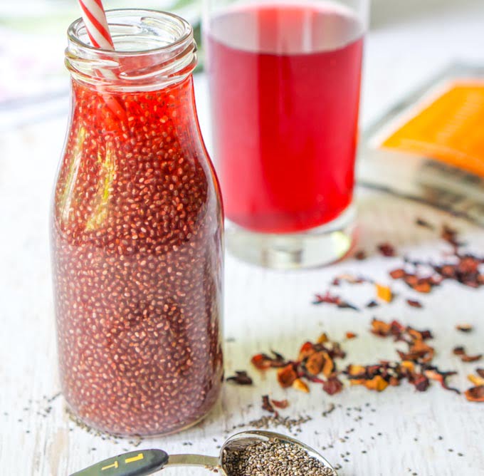 Photo of bottle of red chia seed drink with straw and scattered herbal loose leaf tea and a measuring spoon of chai seeds.