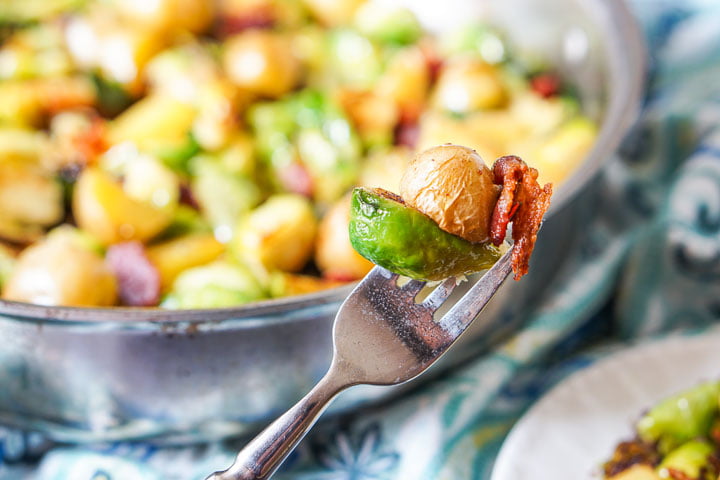 A fork with a Brussels sprout, piece of potato and bacon and pan in background