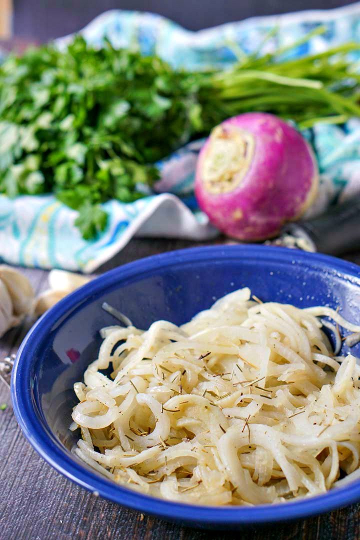closeup of a blue bowl with rosemary & garlic turnip noodles with a raw turnip and parsley in the background