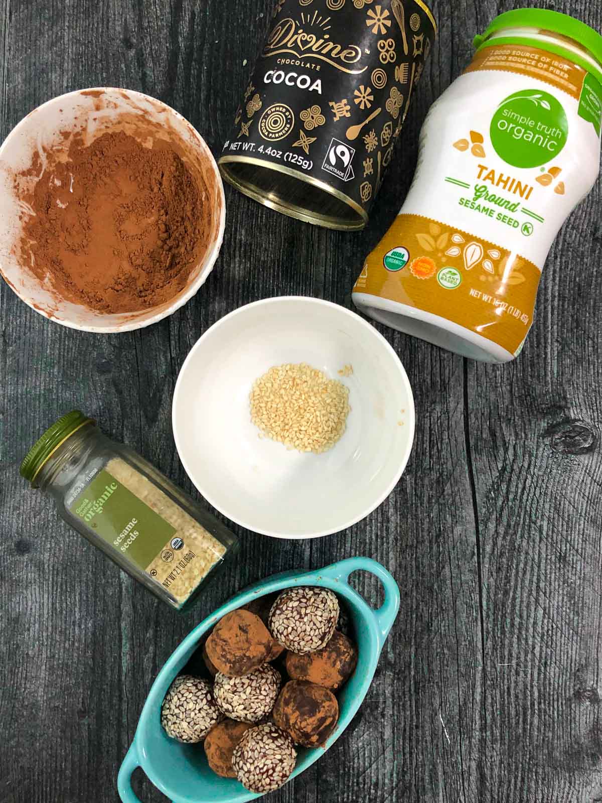 blue bowl with chocolate tahini balls, a bowl of sesames and a bowl of cocoa powder 