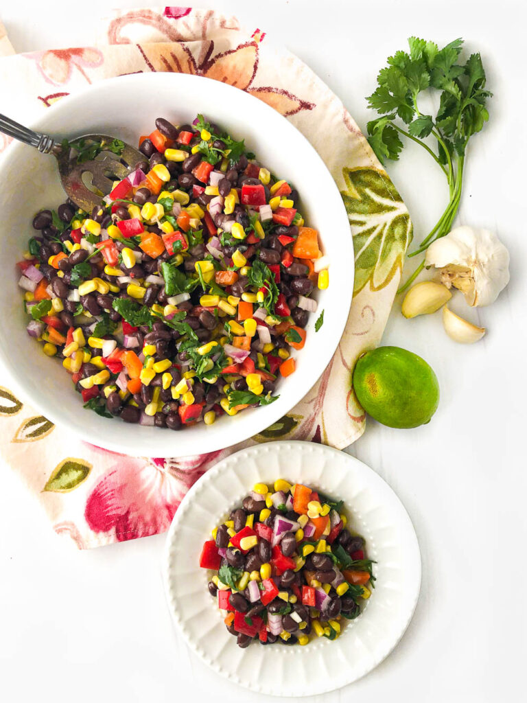 aerial view of white bowl and plate with black bean and corn salad with a lime, garlic and cilantro bunch