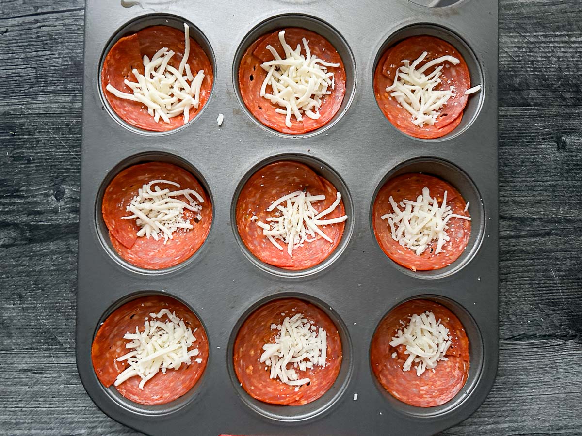 Muffin tin with the uncooked pieces of pepperoni and a pinch of mozzarella cheese ready to bake.
