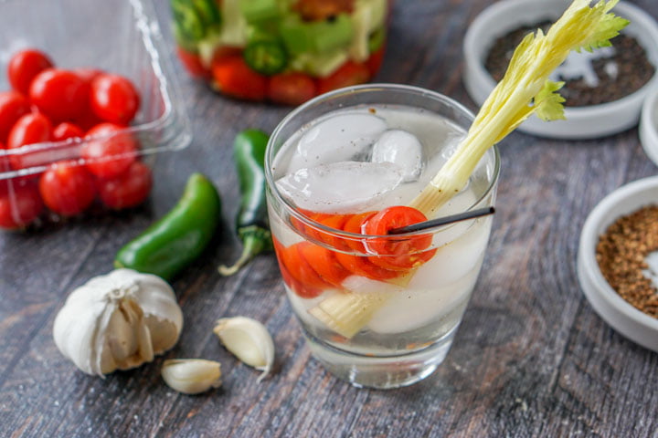 glass with ice, tomato on a stir and celery stalk with garlic, jalapenos and tomatoes in the background