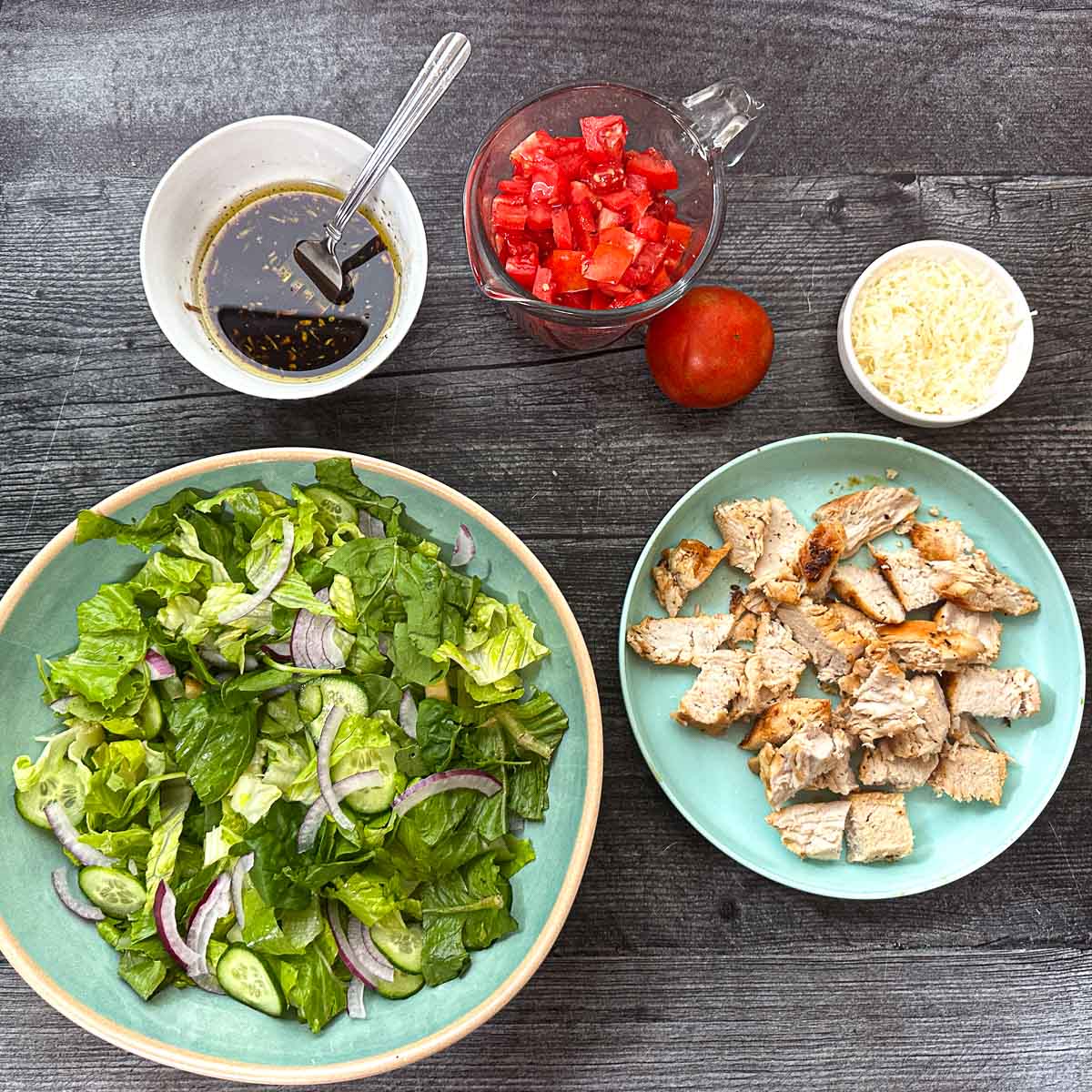 Recipe ingredients - balsamic dressing, a bowl of lettuce, chopped grilled chicken, fresh grated cheese and chopped ripe tomatoes.