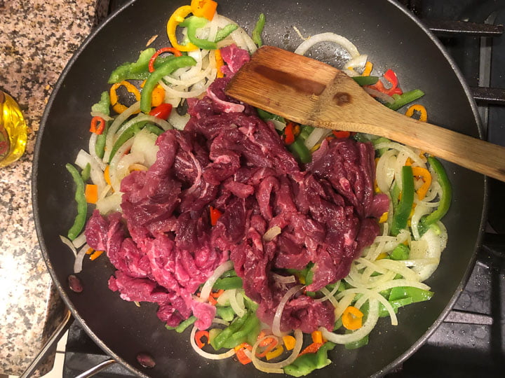 skillet on the stove filled with raw onions, peppers and strips of steak
