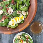 aerial view of wooden bowl and plate with spinach salad and bowl of bacon dressing and text