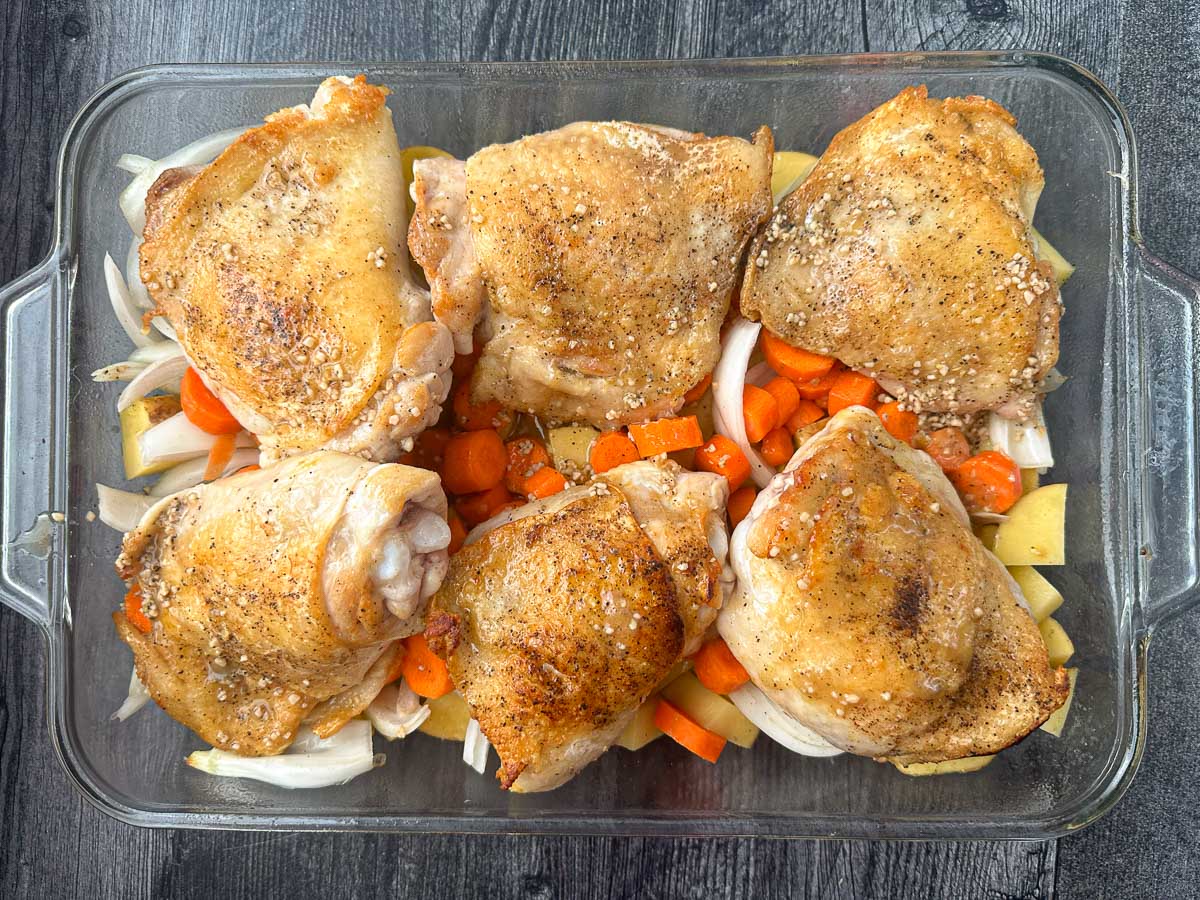 casserole dish with raw veggies and browned chicken thighs ready to bake