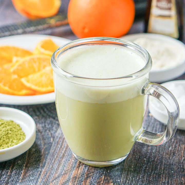 glass mug of low carb orange creamsicle protein matcha drink with bowls of orange slices and matcha in the background