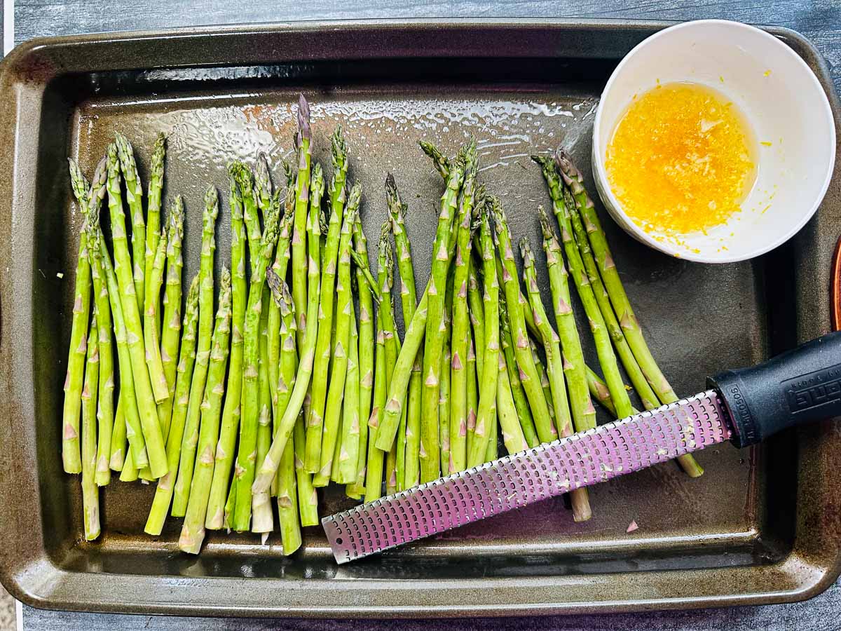 raw asparagus spears on a baking sheet with a bowl of olive oil and crushed garlic