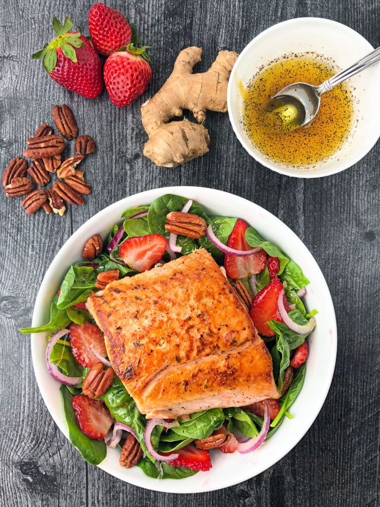 aerial view of salmon spinach salad with a bowl of dressing and fresh strawberries, ginger and pecans