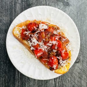 White plate with a piece of savory French toast topped with roasted tomatoes, bacon and sprinkling of parmesan cheese.