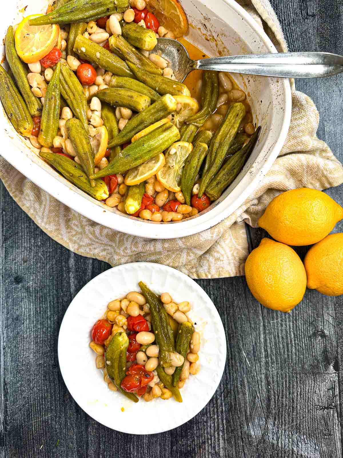 Aerial view of a baking dish and white plate with the okra and tomatoes side dish with fresh lemons.
