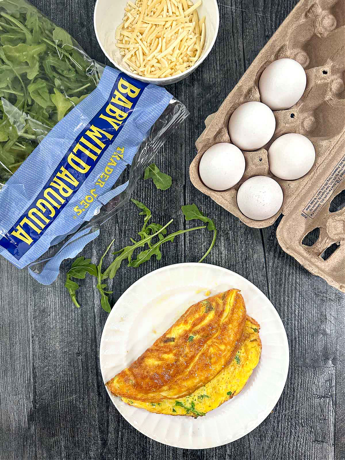 aerial view of arugula omelet on white plate with carton of eggs and a bag of arugula leaves