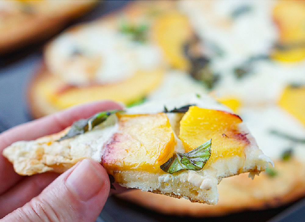 a hand holding a piece of peach caprese pizza with a bite taken
