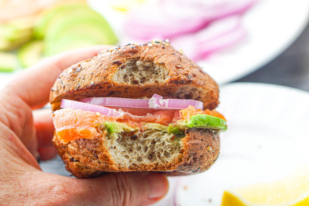 a hand holding a smoked salmon bagel sandwich with a bite taken out