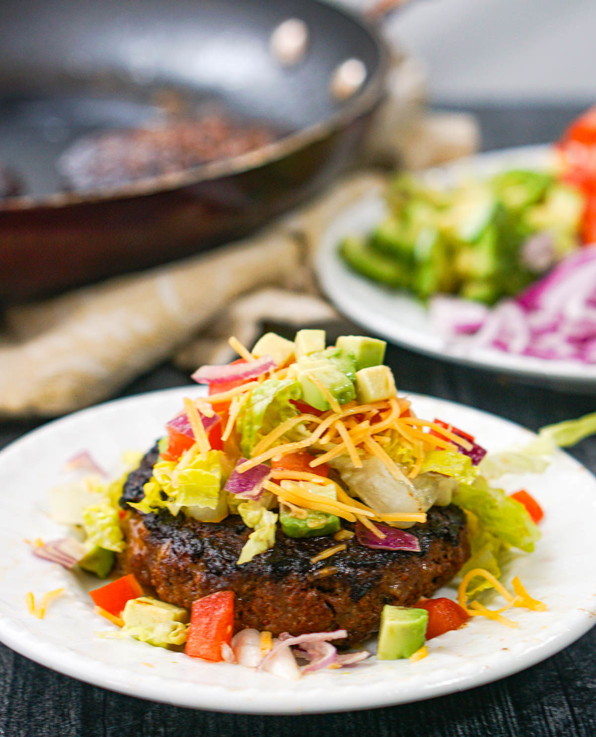 closeup of a keto taco burger with lettuce, onions, avocado and tomatoes on top and a pan and fixing in the background