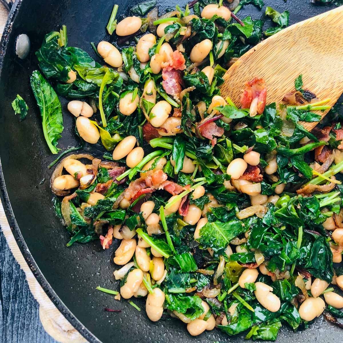 pan with sautéed white beans and greens with bacon and a wooden spoon