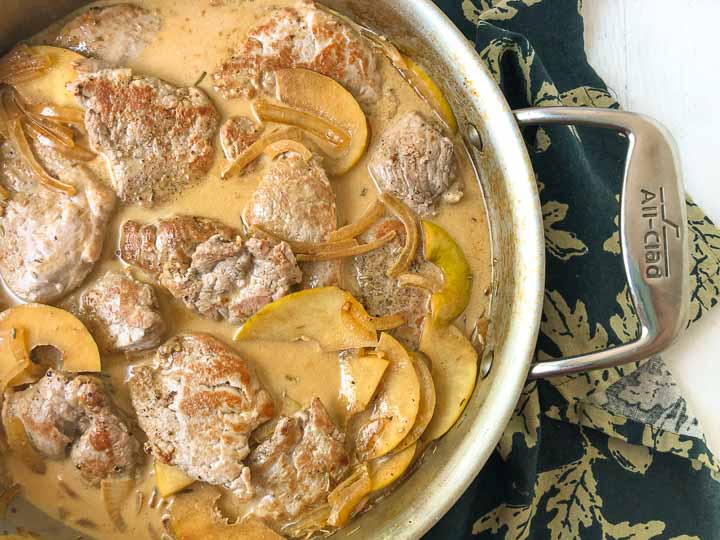 closeup of pan with creamy pork tenderloin and apples on dark greek floral tea towel