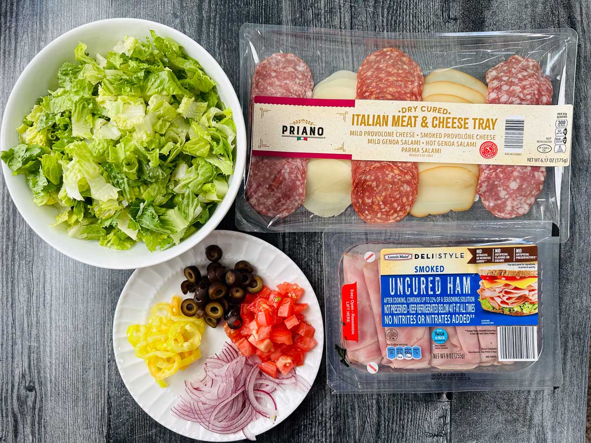 recipe ingredients - bowl of lettuce, meats and cheese, olives, tomatoes, banana pepper rings and red onion slices