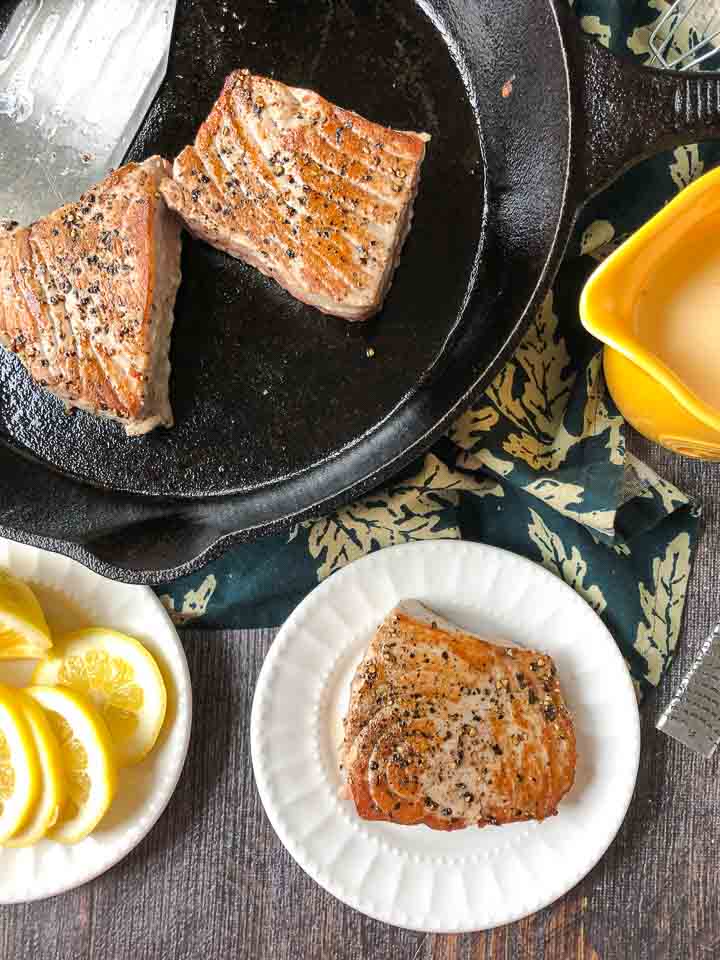 cast iron skillet and a white plate with pepper tuna steaks, sliced lemon on a plate and yellow pitcher with lemon dijon sauce