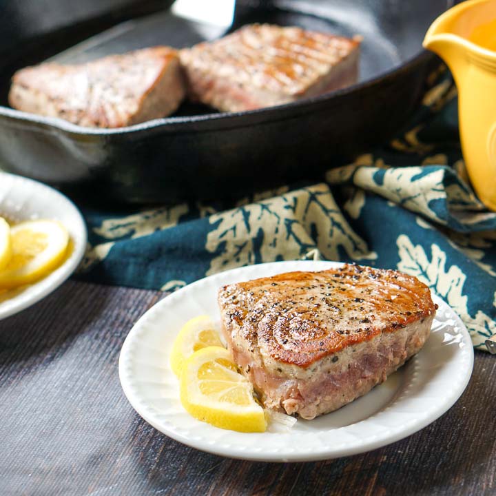 white plate with pepper tuna steak, lemon slices and cast iron skillet in the background