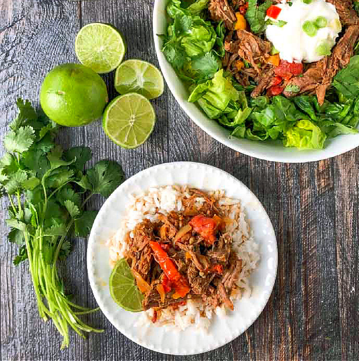 Aerial view of a white plate with a rice topped with the barbacoa beef and fresh limes and cilantro next to it.