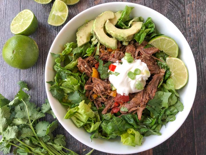 salad bowl with beef, avocado, sour cream and sprigs of cilantro and limes beside it.
