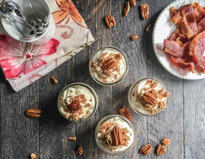 aerial view of 4 jars of no bake cheesecake, spoons and plate of bacon with raw pecans scattered