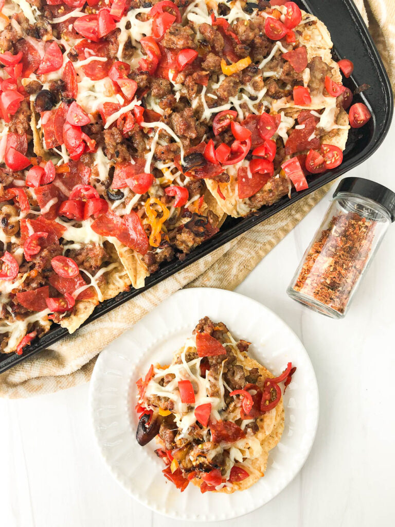 aerial view of a tray of sausage pizza nachos and a white plate with a serving and hot pepper flakes