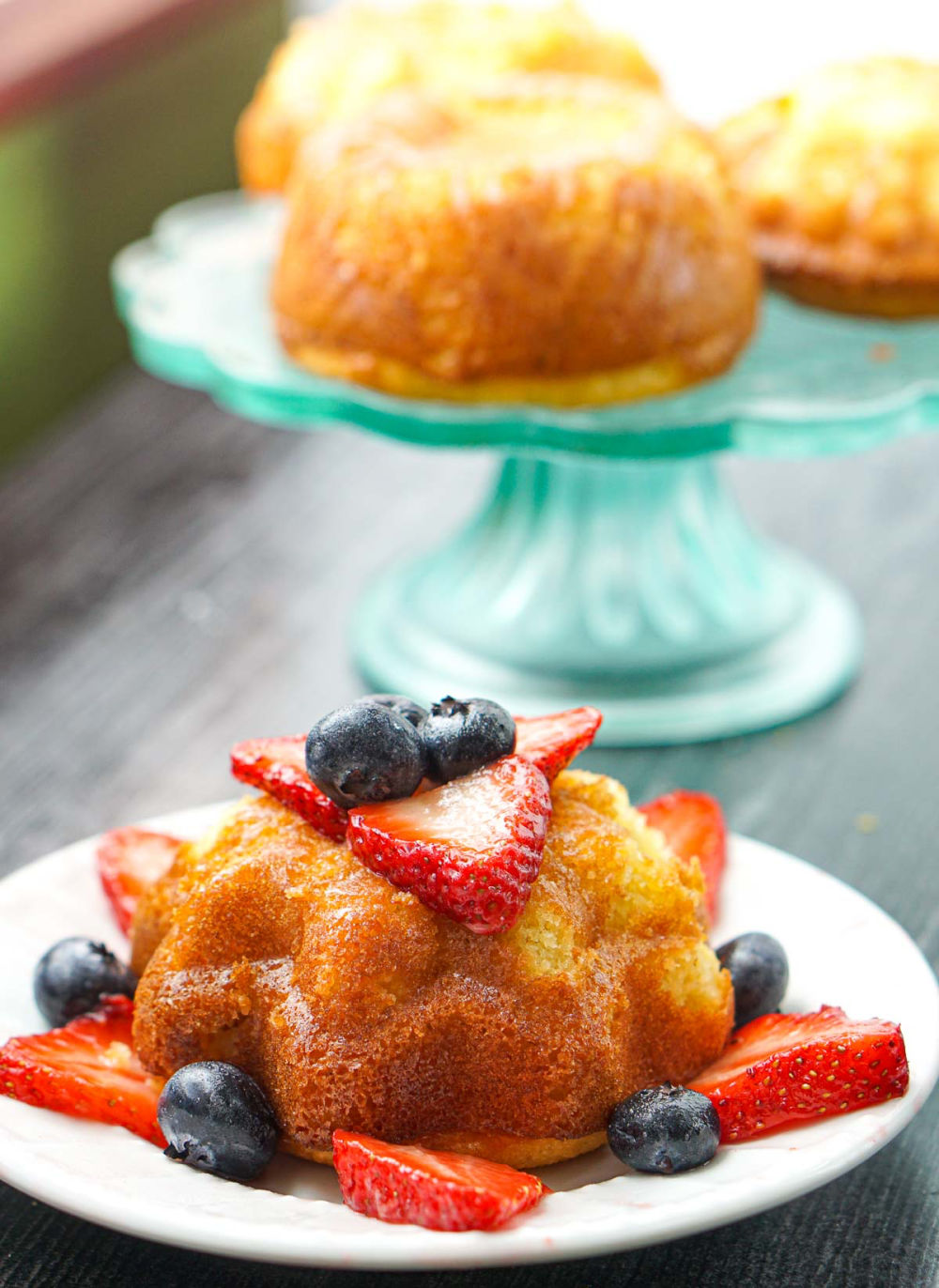 plate with lemon bundt cake with berries and cake stand behind it