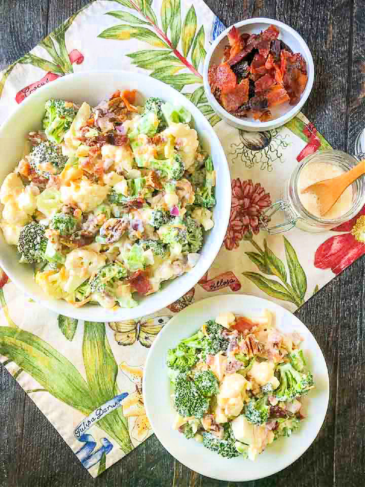aerial view of a bowl and plate with creamy broccoli salad and a bowl of bacon on floral cloth
