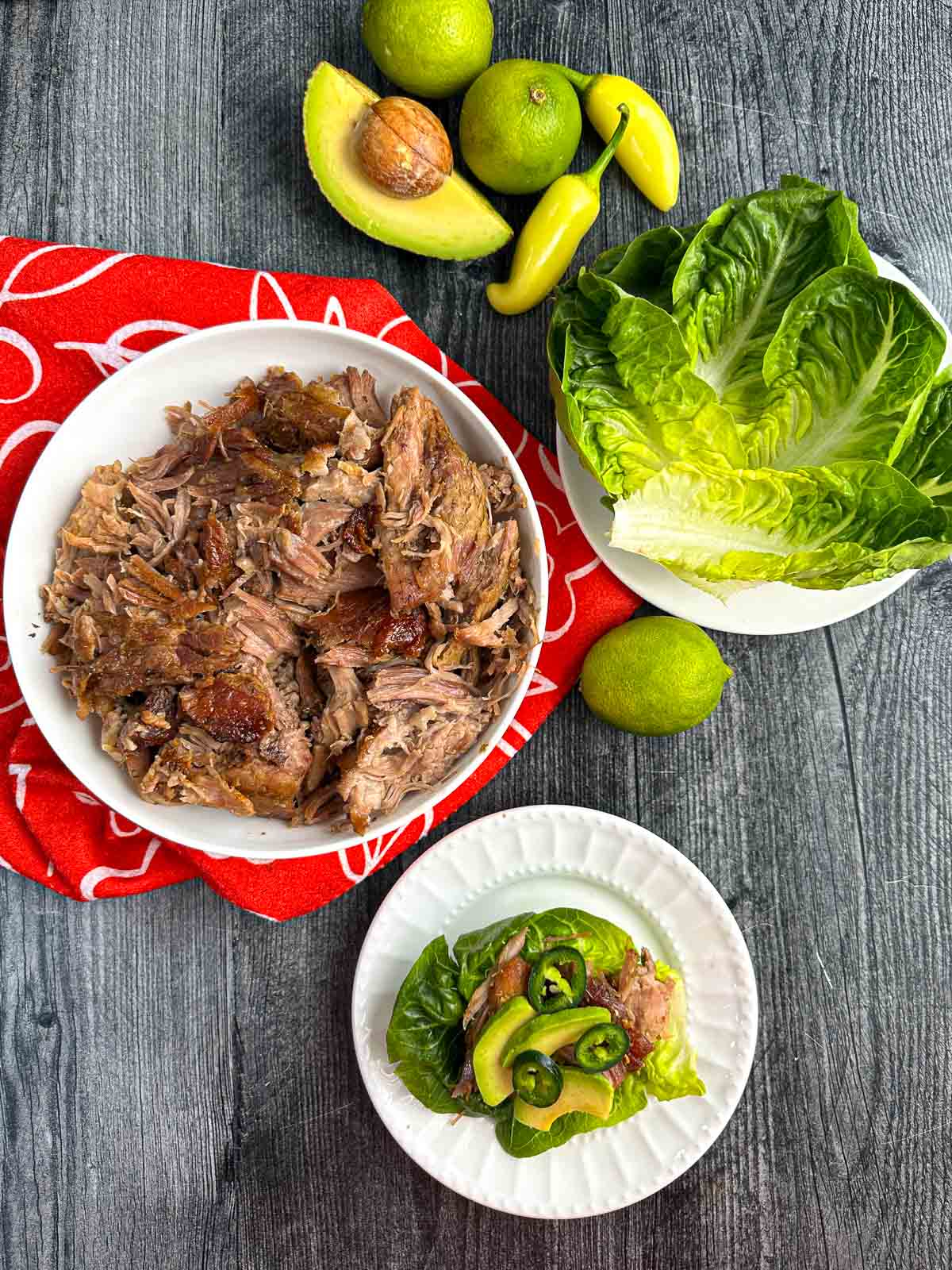Aerial view of a bowl of slow cooker pork carnitas, lettuce wraps and fresh avocados, limes and peppers.