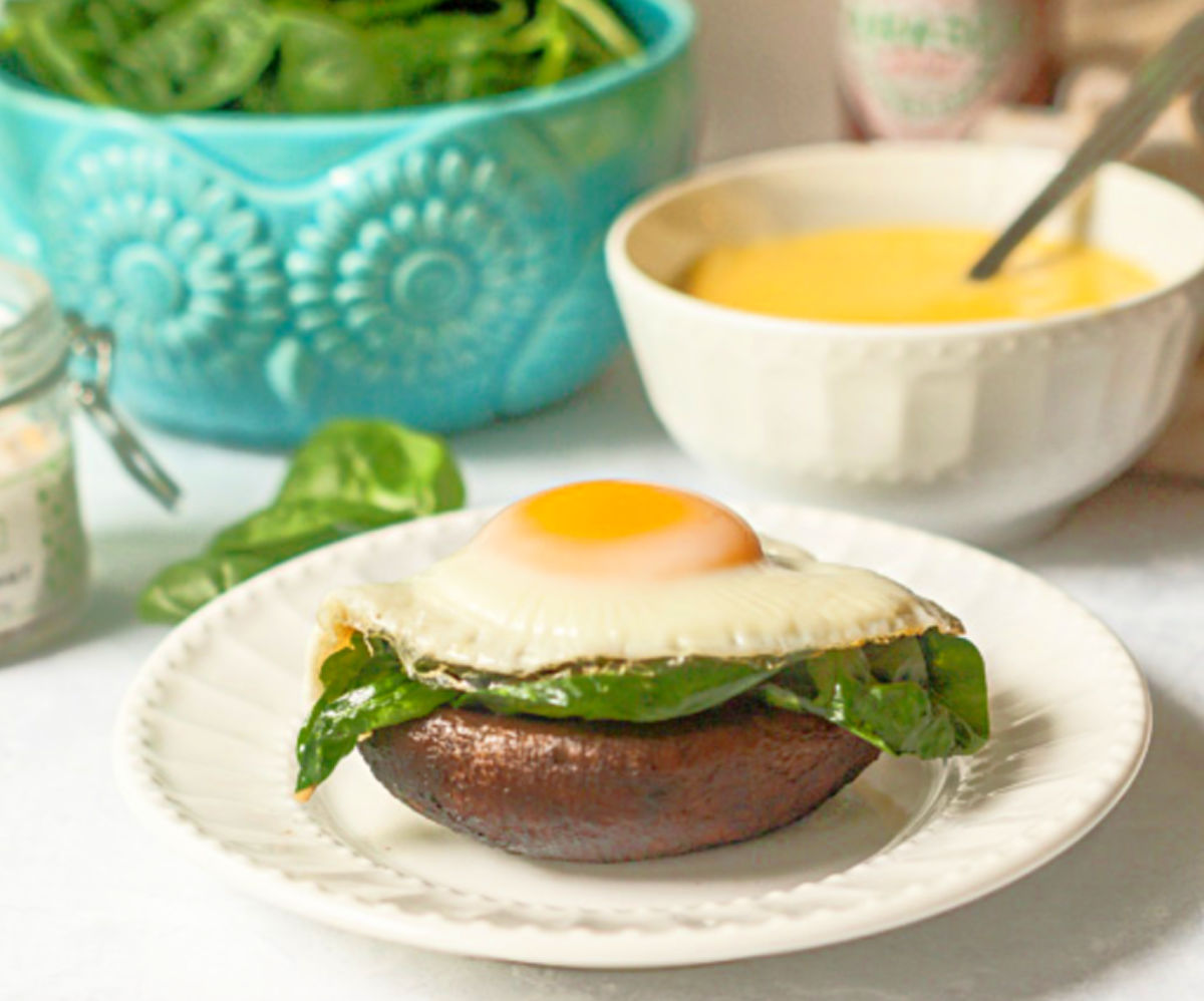 Portobello Eggs with Hollandaise Sauce - low carb breakfast, lunch or dinner! Closeup of a white plate with a portobello mushroom topped with a spinach and an easy over egg with holiday sauce.