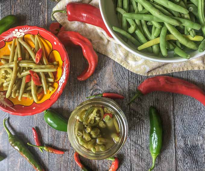 aerial view of a red bowl and jar with pickled green beans and a bowl of raw green beans and raw hot peppers