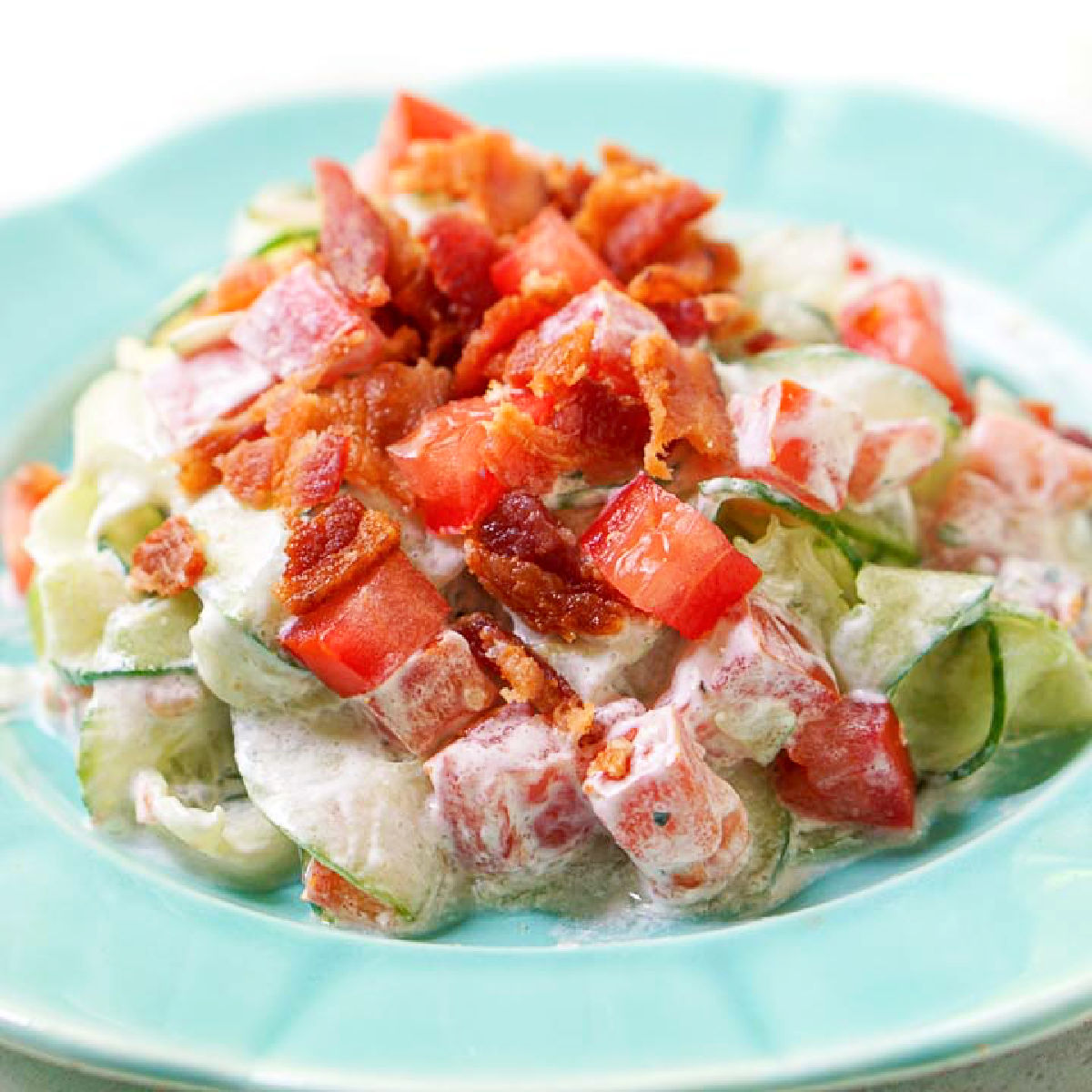 Closeup of an aqua plate with a serving of creamy cucumber salad with tomatoes, ranch and bacon.