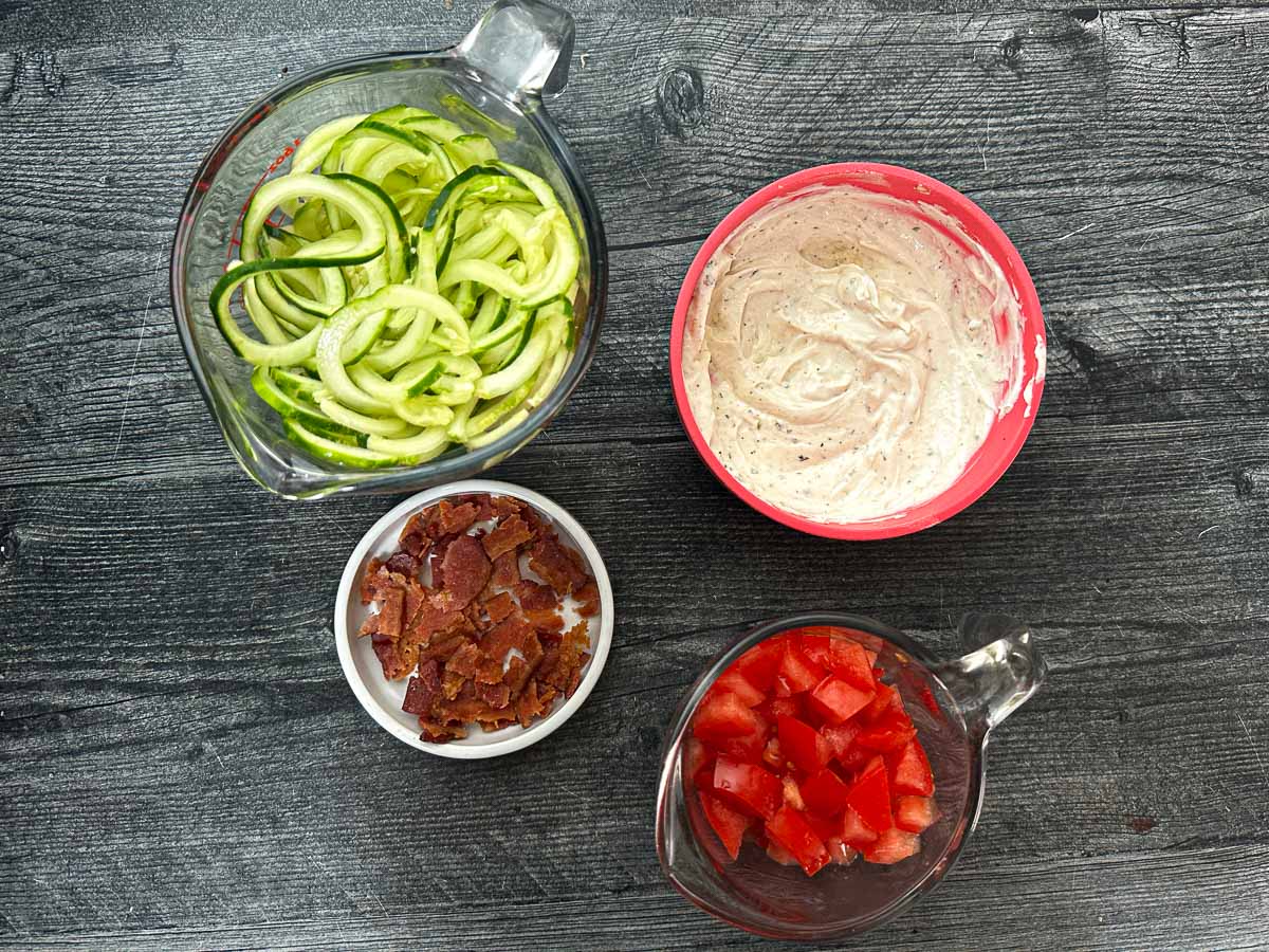 Recipe ingredients - ranch made with sour cream, bacon bits, cucumber noodles and chopped tomatoes.