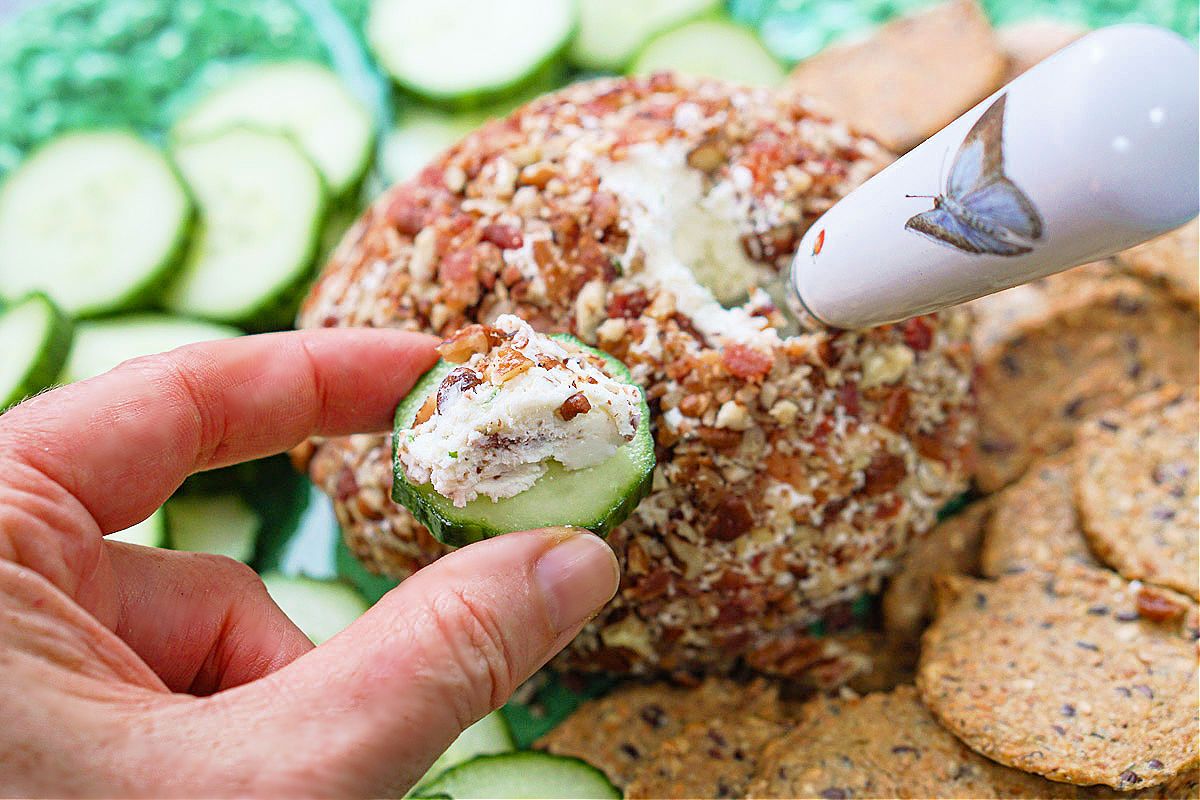 fingers holding a slice of cucumber with cheese spread on