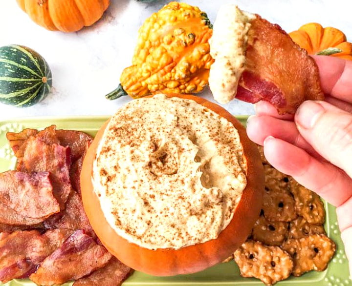 hand with a bacon chip dipped into the pumpkin cheesecake dip and the platter in the background