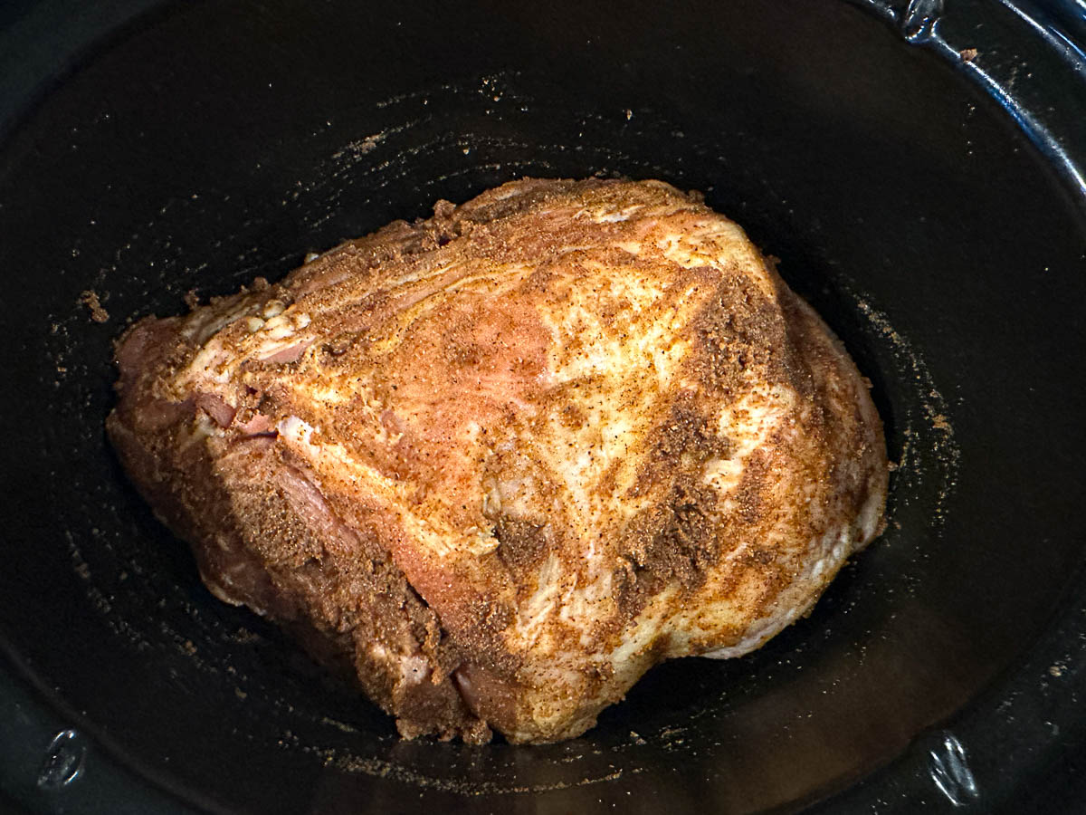 Crock pot with a raw pork roast and a spice rub.