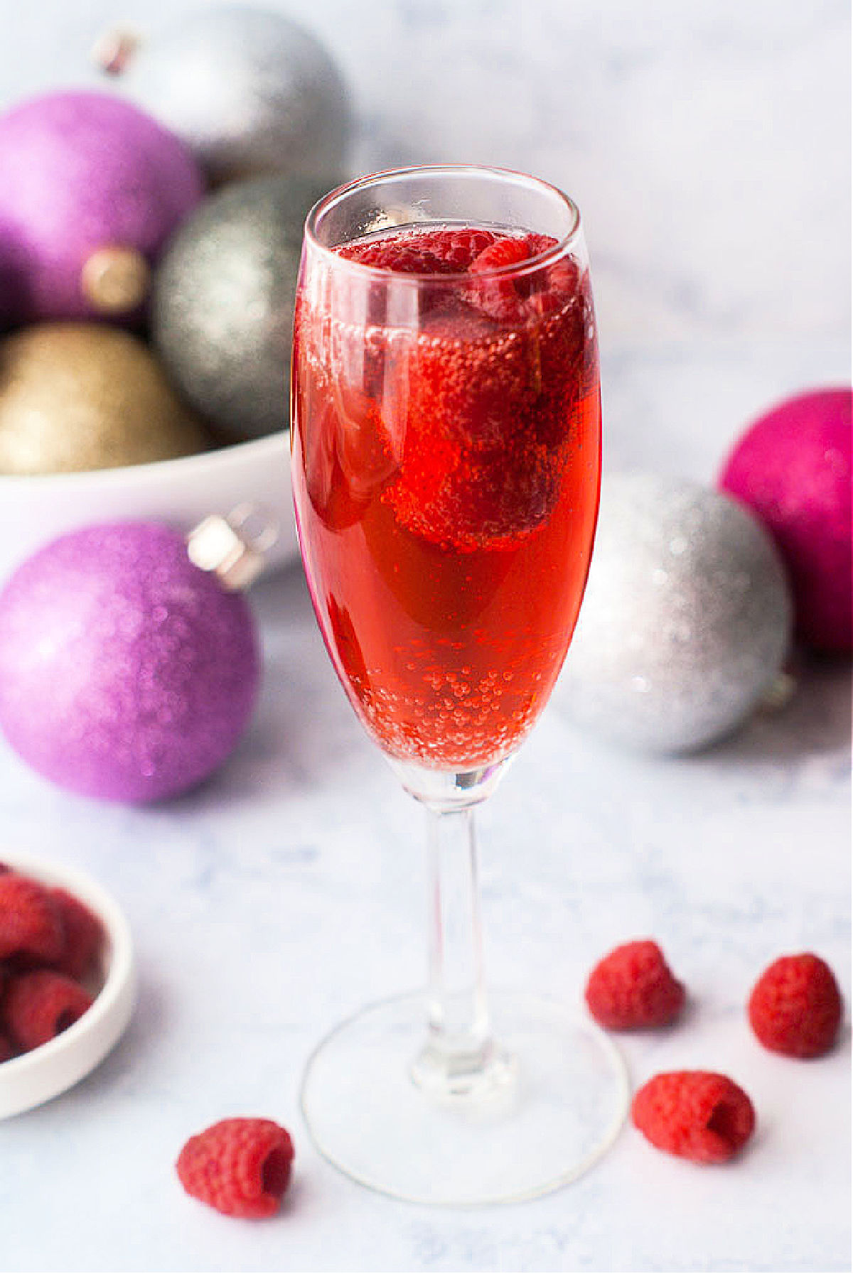 closeup of a champagne glass of raspberry mimosa drink with fresh raspberries and Christmas bulbs in the background