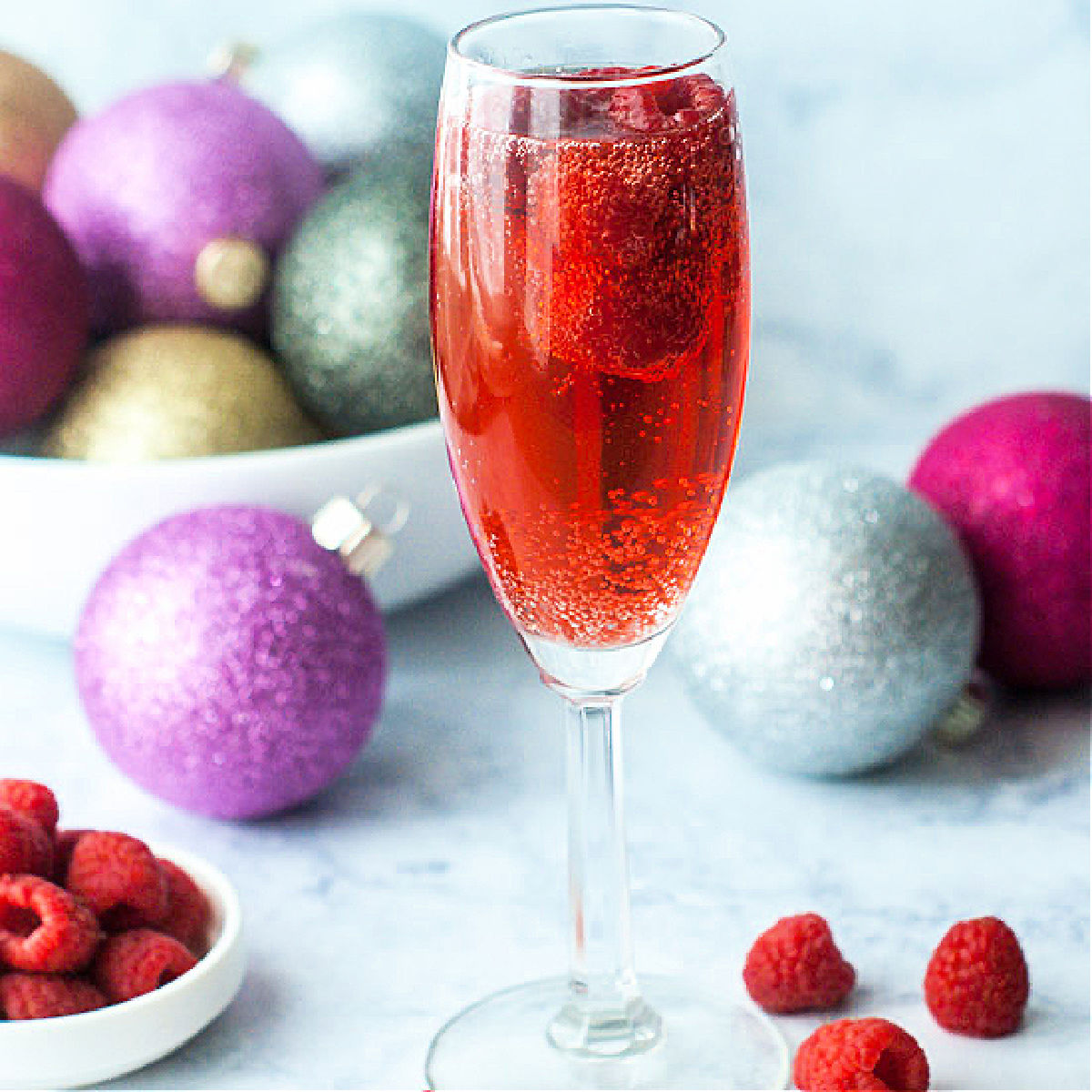 glass of raspberry mimosa drink with fresh raspberries and Christmas bulbs in the background