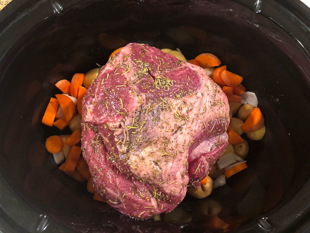 black slow cooker crock with raw veggies and a raw lamb roast with an herb rub all over