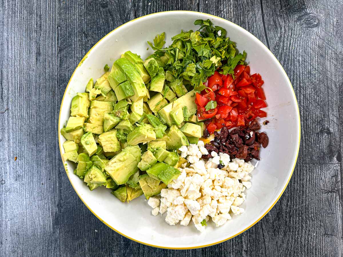 White bowl with chopped avocados, cilantro, olives, tomatoes and feta cheese.