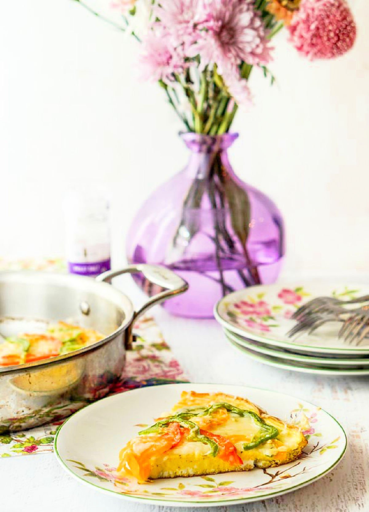 A tall photo of a table setting with a slice of the pepper gruyere frittata with a purple vase with fresh flowers in the background.