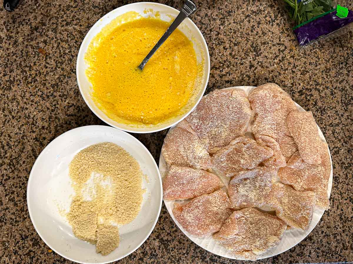 Breading the cutlets with bowls of almond flour, egg batter and raw chicken pieces on a plate.