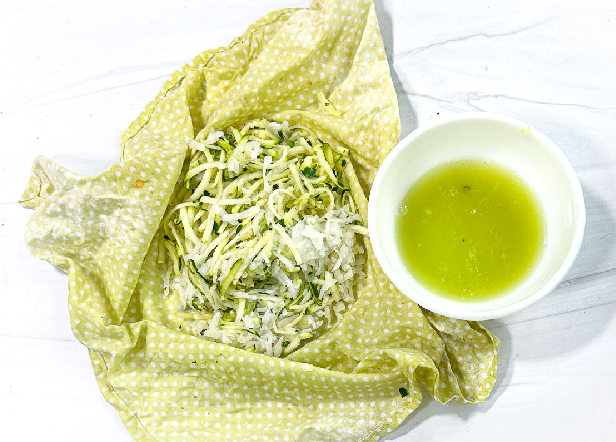 Green cloth with shredded zucchini and onion and bowl of liquid from them.