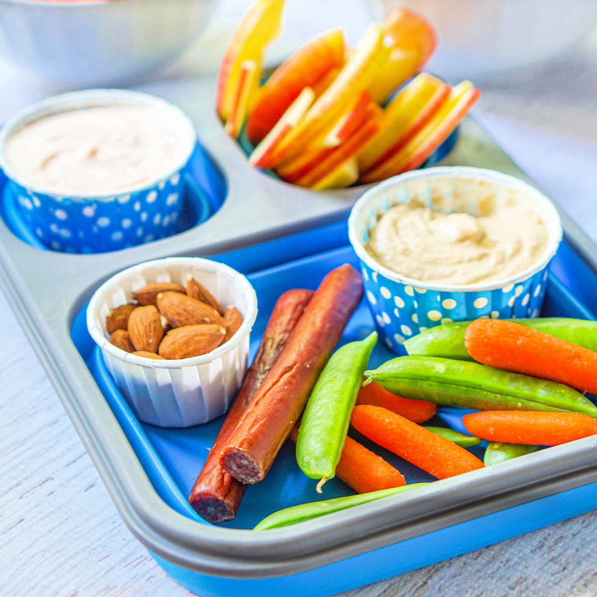 blue lunch box with hummus, almonds, beef sticks, veggies and apples