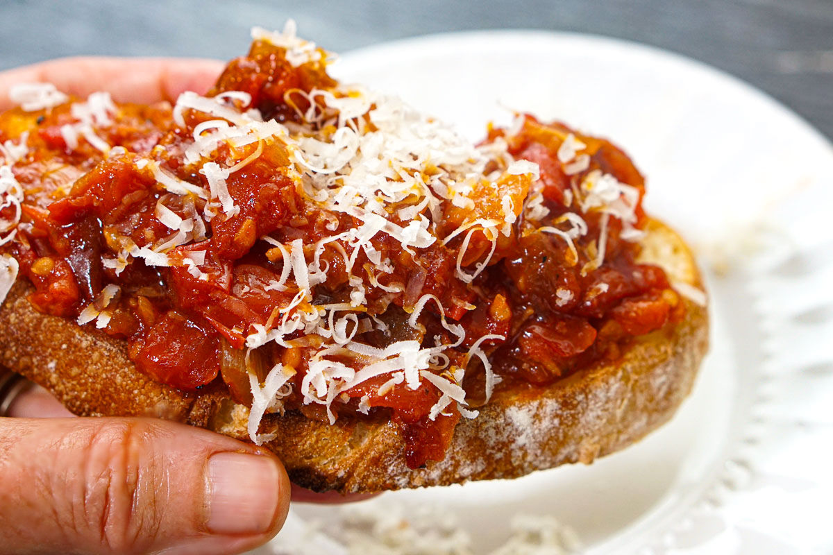 Hand holding a piece of toasted crusty bread with the finished tomato chutney on top and grated cheese.