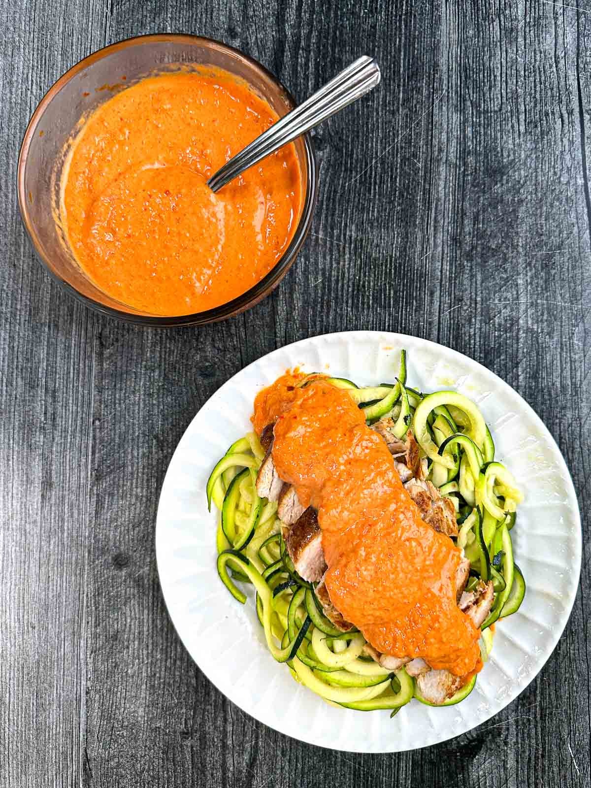 Aerial view of a bowl with creamy red pepper sauce and a white plate with zucchini noodles and chicken breast covered with red pepper sauce.